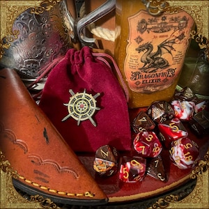 May include: A maroon velvet drawstring pouch featuring a gold and black compass pin with crossed swords. Beside it sits a bottle labeled Dragonfire Elixir with a dragon illustration, a brown leather sheath, and several multi-sided gaming dice.