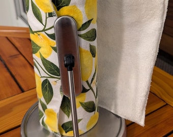 May include: A stainless steel paper towel holder with a lemon-patterned paper towel roll and a white cloth towel hanging from it. The holder is on a wooden surface.