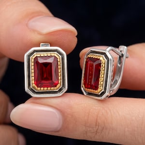 Pode incluir: Um par de brincos com pedras retangulares facetadas em vermelho profundo, engastadas em biséis dourados com bordas texturizadas. As pedras são emolduradas por armações octogonais prateadas oxidadas e possuem fechos de alavanca articulados.