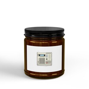 May include: A brown glass candle jar with a black lid, featuring a white rectangular label on the front. The label displays a detailed illustration of a light gray music production sampler with a digital screen, various buttons, and a grid pad.