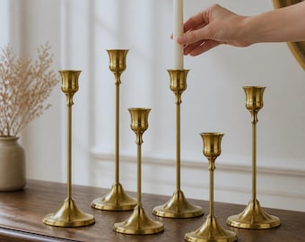 May include: A set of six gold-toned metal candle holders with slender stems and flared bases. Each holder features a cup-shaped top designed to secure a taper candle. One white taper candle is positioned in the tallest central candle holder.