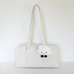 Puede incluir: Un bolso tote rectangular de tela a rayas beige y blanca con dos asas largas para el hombro. Un pequeño dije de peluche blanco con forma de nube, que presenta una cara sonriente y dos pies colgantes de color marrón oscuro, está sujeto al frente.