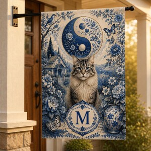 Puede incluir: Una bandera de jardín rectangular con un diseño azul y blanco de estilo chinoiserie. El estampado muestra un gato atigrado central, un símbolo yin-yang, una casa de campo, motivos florales, mariposas y un jarrón. Un emblema circular contiene la letra M.