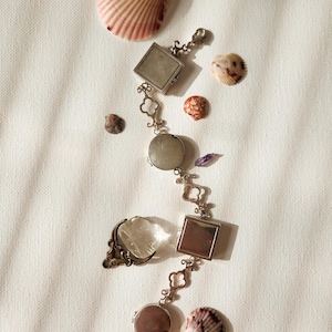 May include: A silver-toned metal charm bracelet featuring alternating square and circular locket-style pendants connected by clover-shaped links. A clear, spherical glass charm with a decorative metal base is positioned next to the bracelet.