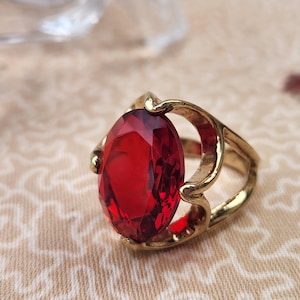 May include: A gold-toned ring featuring a large, oval-cut, deep red faceted gemstone set in a decorative metal band with open, curved side details that frame the central stone, creating a bold and reflective appearance on the surface.