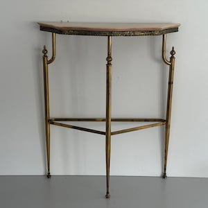 May include: A semi-circular console table featuring a light-colored stone top with a decorative gold-toned metal edge. The base consists of three slender gold-toned metal legs connected by a triangular support frame with ornate finial accents.