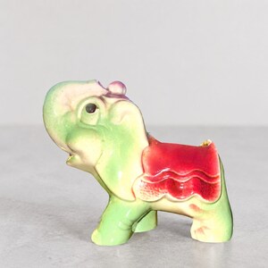 Peut inclure: Une figurine d'éléphant en céramique au corps vert clair et crème avec une finition brillante. L'éléphant a la trompe levée et est orné d'une couverture de selle rouge vif drapée sur son dos, présentant des bords festonnés détaillés.