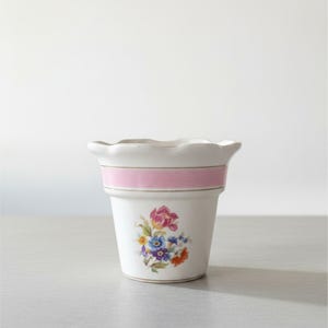 May include: A white ceramic planter with a scalloped rim and a horizontal pink band encircling the upper section. The body features a central floral decal displaying a cluster of colourful flowers including pink, blue, yellow, and orange blossoms.