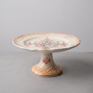 May include: A ceramic cake stand featuring a scalloped edge with a peach-colored rim. The center of the plate and the base are decorated with a floral pattern of pink roses and blue accents, complemented by delicate gold scrollwork details.
