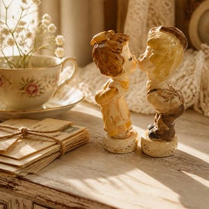 May include: Two ceramic figurines of a young girl and a young boy standing on circular bases, leaning toward each other as if to kiss. The girl has brown hair and a light-colored dress, while the boy has light hair, a cap, and dark trousers.
