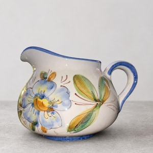 May include: A ceramic creamer pitcher with a cream-colored base, featuring hand-painted blue floral motifs and green leaf accents. The pitcher has a curved handle, a pouring spout, and a blue painted rim that matches the handle and base trim.