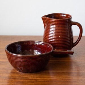 May include: A dark brown ceramic pitcher and a matching bowl with a handle. Both pieces feature a glossy, glazed finish with a distinct horizontal ribbed texture covering the exterior surfaces, showcasing a deep, rich, and uniform brown color.