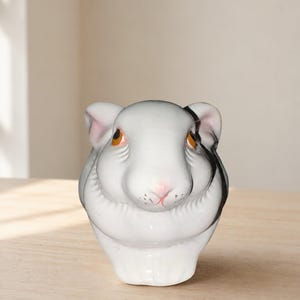 May include: A ceramic figurine shaped like a guinea pig head, featuring a white base color with black markings around the ears and eyes. The animal has orange-brown eyes, a small pink nose, and detailed whiskers painted on its snout.