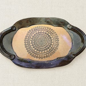 May include: An oval ceramic tray featuring a light tan center with a dark brown, intricate, circular lace-like pattern. The tray has a wide, dark, glossy rim with a metallic sheen and two integrated handles on the left and right sides.