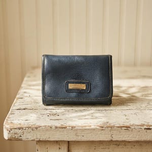 May include: A dark navy blue bi-fold wallet with a textured surface and a rectangular gold-toned metal plaque on the front flap. The plaque features the embossed text Liz Claiborne. The wallet has visible stitching along the edges.