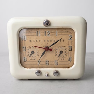 May include: A vintage Westinghouse rectangular table clock with an off-white plastic casing. The clock face is tan with black numerals, featuring a central time display and two smaller sub-dials labeled START and STOP. It has three silver knobs.