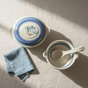 May include: A ceramic lidded casserole dish with blue concentric rings and a floral motif, a matching open bowl with a blue-rimmed interior, a ceramic spoon, and a folded light blue linen napkin. The set features a cream-colored base finish.