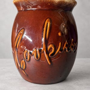 May include: A glossy, dark brown ceramic cookie jar with a rounded body and a flared rim. The word Cookies is embossed in a raised, cursive script across the front. The top edge features a lighter, cream-colored drip glaze effect.