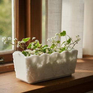 May include: A rectangular white milk glass planter featuring a scalloped top edge and an embossed grape and vine pattern on the exterior. The planter is filled with green leafy vines and small white flowers, creating a decorative display.