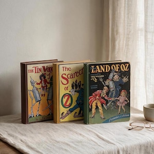 May include: Three vintage hardcover books titled The Tin Woodman of Oz, The Scarecrow of Oz, and The Land of Oz. Each book features colorful, detailed illustrations on the front cover depicting characters from the Oz series by L. Frank Baum.