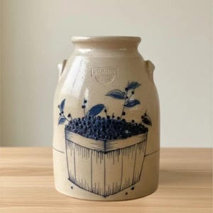 May include: A cream-colored ceramic crock with two side handles, featuring a blue hand-painted illustration of a wooden basket overflowing with berries. The design includes a small stamped logo near the rim and a speckled texture throughout.