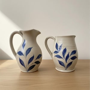 May include: Two off-white ceramic pitchers featuring a hand-painted blue leaf pattern. The left pitcher has a curved spout and a rounded body, while the right pitcher has a straighter rim and a slightly more cylindrical shape, both with handles.