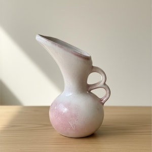 May include: A ceramic pitcher with a rounded base and a tall, flared spout. The vessel features a smooth, matte finish in a soft off-white color with subtle, blended pink gradients. It includes a distinctive handle with two stacked circular loops.
