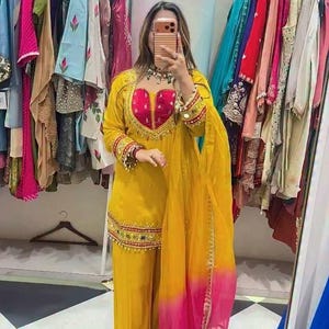 Può includere: Un completo a tre pezzi giallo brillante composto da una tunica a maniche lunghe con scollo a cuore rosa scuro, pantaloni larghi abbinati e un dupatta giallo trasparente con orlo sfumato rosa, decorato con ricami dorati e piccoli specchi.