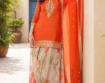 May include: A bright orange long-sleeved tunic featuring intricate gold embroidery around the neckline and hem, paired with a flowing, floor-length skirt adorned with a multi-coloured floral print and a matching orange sheer dupatta with gold trim.
