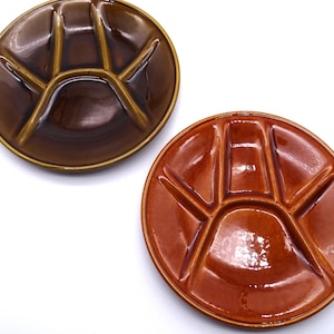 May include: Two round ceramic plates with a glossy finish, each featuring a raised, star-shaped divider pattern in the centre. One plate is a deep dark brown, while the other is a lighter, warm terracotta orange colour with a smooth, reflective surface.