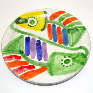May include: A round ceramic plate with a speckled off-white base featuring hand-painted abstract designs. The artwork includes two large green leaf-like shapes with dotted centres, accented by vibrant stripes in yellow, orange, blue, and purple.