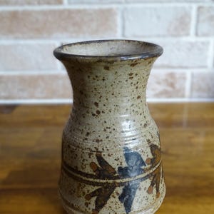 May include: A stoneware vase with a speckled beige and tan finish, featuring a flared rim and a tapered neck. The lower body is decorated with a horizontal band containing dark brown and black abstract leaf-like motifs painted on the surface.