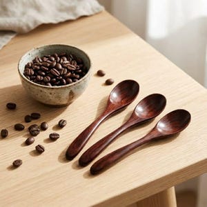Puede incluir: Tres cucharas de madera de color marrón oscuro con cuencos lisos y redondeados y mangos curvos dispuestos en diagonal. Junto a las cucharas hay un cuenco de cerámica claro y moteado lleno de granos de café tostado, con varios granos sueltos cerca.
