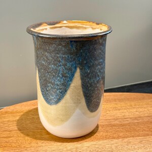 May include: A cylindrical ceramic vase featuring a layered glaze design. The top section is a deep, textured blue that drips down into a smooth, light beige middle, transitioning to a solid white base. The rim is dark and defined.
