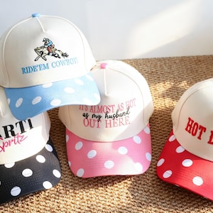 Peut inclure: Quatre casquettes de baseball avec des calottes blanches et des visières colorées à pois blancs. Les casquettes présentent des textes brodés : RIDE'EM COWBOY, SARTI Spritz, IT'S ALMOST AS HOT as my husband OUT HERE, et HOT DAMN en diverses couleurs.