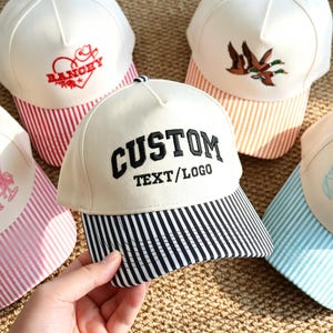 May include: Five baseball caps with white crowns and striped brims. The caps feature various embroidered designs including a red heart with the text RANCHY, flying ducks, the text Birdies & Dirties, the text CUSTOM TEXT/LOGO, and blue seashells.