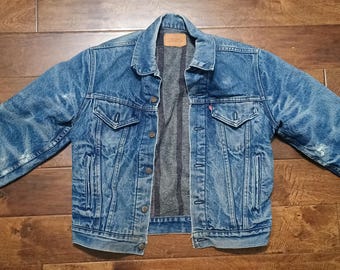 May include: A blue denim jacket with a faded wash, featuring a pointed collar, metal button closures, two chest pockets with buttoned flaps, and two side slit pockets. The interior is lined with a dark grey fabric and includes a leather neck label.