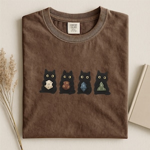 Op de afbeelding: Een bruin t-shirt met ronde hals en een afbeelding van vier zwarte katten, die elk een andere veelzijdige dobbelsteen vasthouden. Het shirt heeft een Comfort Colors merklabel aan de binnenkant van de halslijn. De katten hebben grote gele ogen.