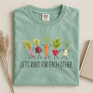 May include: A sage green crewneck t-shirt featuring a graphic of five anthropomorphic vegetables with smiling faces. Below the vegetables, the text reads LETS ROOT FOR EACH OTHER. The shirt has a Comfort Colors brand label at the inner neckline.