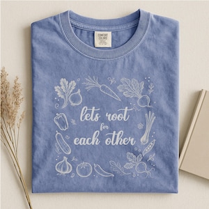 Peut inclure: Un t-shirt bleu à col rond avec un motif graphique en lignes blanches représentant divers légumes entourant le texte "lets root for each other". Le t-shirt présente une étiquette de marque Comfort Colors à l'intérieur du col.