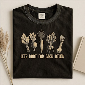 Peut inclure: Un t-shirt noir à col rond avec une illustration graphique beige représentant cinq légumes racines différents alignés. Sous les légumes, le texte LETS ROOT FOR EACH OTHER est inscrit dans une police sans empattement en majuscules.