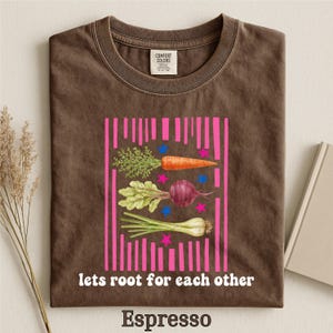 May include: A brown t-shirt featuring a graphic of a carrot, a beet, and a parsnip arranged vertically. The design includes pink vertical stripes, small blue and pink stars, and the white text "lets root for each other" printed below the vegetables.