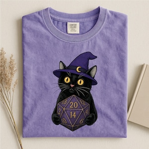 May include: A lavender short-sleeve t-shirt featuring a graphic of a black cat wearing a purple witch hat with a yellow crescent moon. The cat is holding a purple twenty-sided die with gold numbers. The shirt has a Comfort Colors brand label.