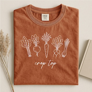 May include: A rust-colored short-sleeve t-shirt featuring a white line-art graphic of various vegetables including a carrot and leafy greens. The text "crop top" is printed in white script below the vegetables. A Comfort Colors brand label is visible.