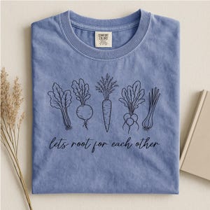 Könnte beinhalten: Ein kornblumenblaues T-Shirt mit Rundhalsausschnitt und einer schwarzen Strichzeichnung verschiedener Wurzelgemüse, darunter eine Karotte und ein Rettich. Unter dem Gemüse steht der Text "lets root for each other" in Schreibschrift.