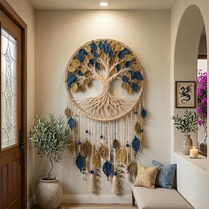 May include: A circular macrame wall hanging featuring a tree of life design with cream-coloured cord, accented by numerous blue and gold crocheted leaves. The piece includes hanging strands with additional leaves, tassels, and blue beads.
