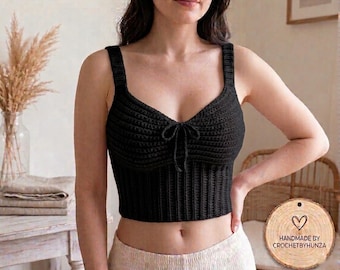 May include: A black crochet crop top featuring a V-neckline with a central tie detail and wide shoulder straps. The bodice has a horizontal ribbed texture, while the lower section features a vertical ribbed pattern. A wooden tag reads HANDMADE BY CROCHETBYHUNZA.
