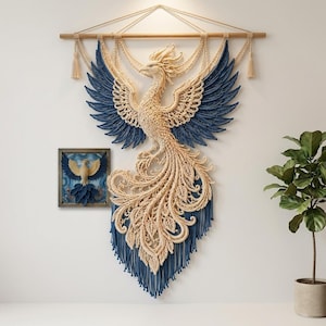 May include: A large macrame wall hanging featuring a cream and navy blue phoenix with intricate knotted wings and a long, flowing tail, suspended from a wooden rod. Beside it is a square framed artwork depicting a similar phoenix design.