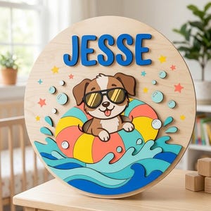 Puede incluir: Un letrero circular de madera para pared con el nombre JESSE en letras azules. El diseño incluye un cachorro marrón y blanco con gafas de sol negras dentro de un flotador a rayas, rodeado de olas de agua azules, burbujas y estrellas.