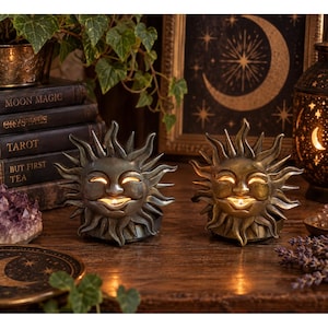 May include: Two sun-shaped candle holders with metallic bronze finishes and smiling faces. Each holder features radiating sunbeams and a central opening where a small lit candle glows from within, casting warm light through the eyes and mouth.